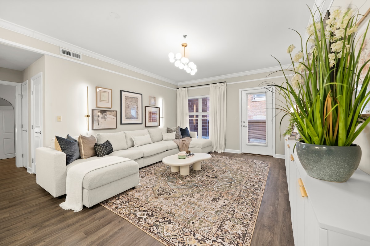 A spacious living area is presented with a light-colored sectional sofa positioned around a low coffee table. Decorative pillows add texture. A large area rug anchors the seating arrangement, while a vibrant plant in a ceramic pot introduces greenery. Natural light is welcomed through double doors.