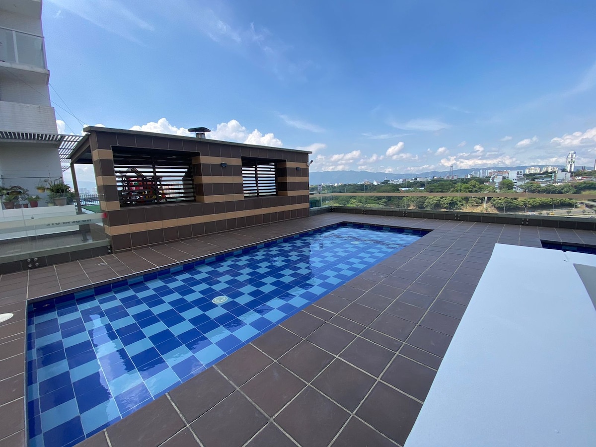 An outdoor pool is framed by modern tiles in a blue and brown pattern. A shaded seating area is visible nearby, with a panoramic view of the surrounding landscape. The sky is clear with scattered clouds, creating a bright ambiance.