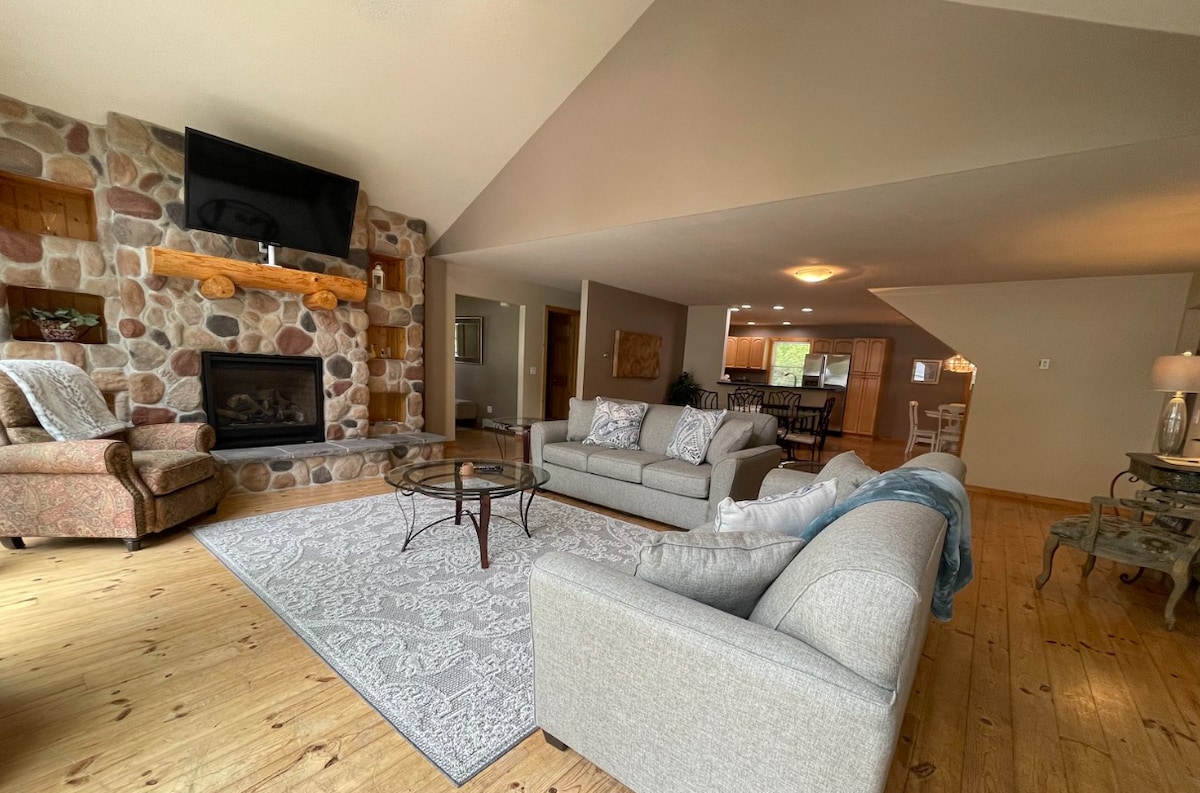 The living area features a cultured stone fireplace with a rustic timber mantel. Two sofas and an armchair are arranged around a glass coffee table atop a textured area rug. Large windows allow natural light to fill the space, contributing to an airy feel.