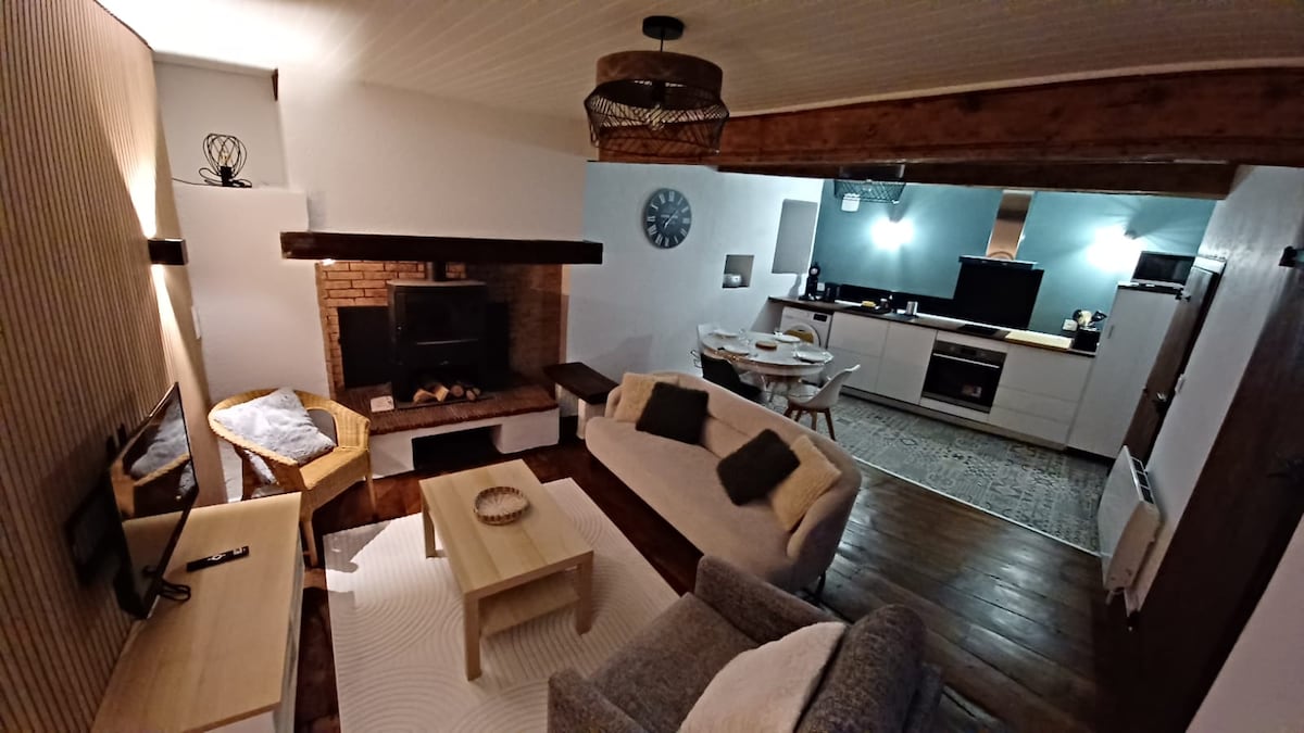 The living space is characterized by a blend of natural wood accents and modern furnishings. A comfortable seating area includes a light-colored sofa and armchair, positioned around a wooden coffee table. A brick fireplace serves as a focal point, with an adjacent dining area visible in the background.