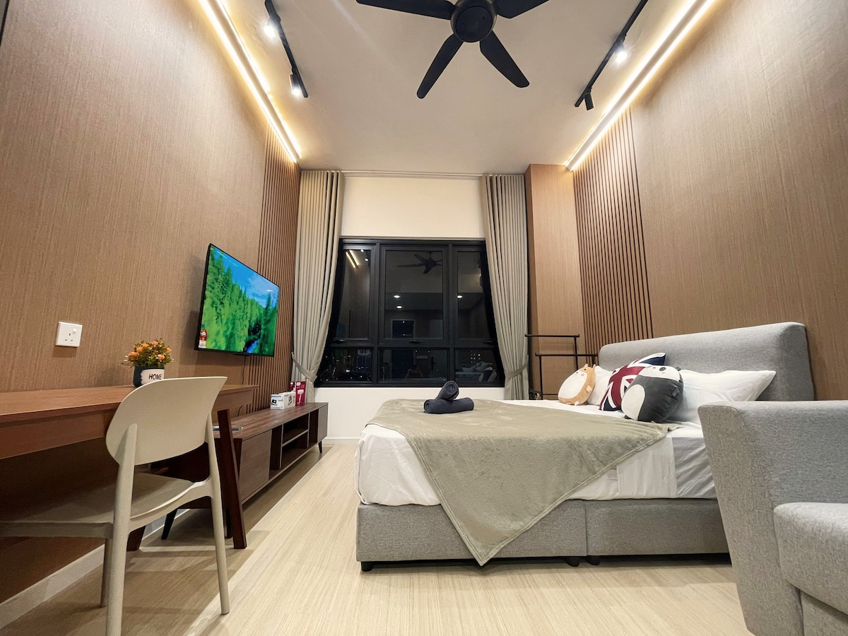 A studio space is showcased with a spacious bed draped in neutral bedding. A comfortable seating area is present alongside a writing desk. A 50-inch TV is mounted on the wall, and large windows allow natural light to illuminate the room.