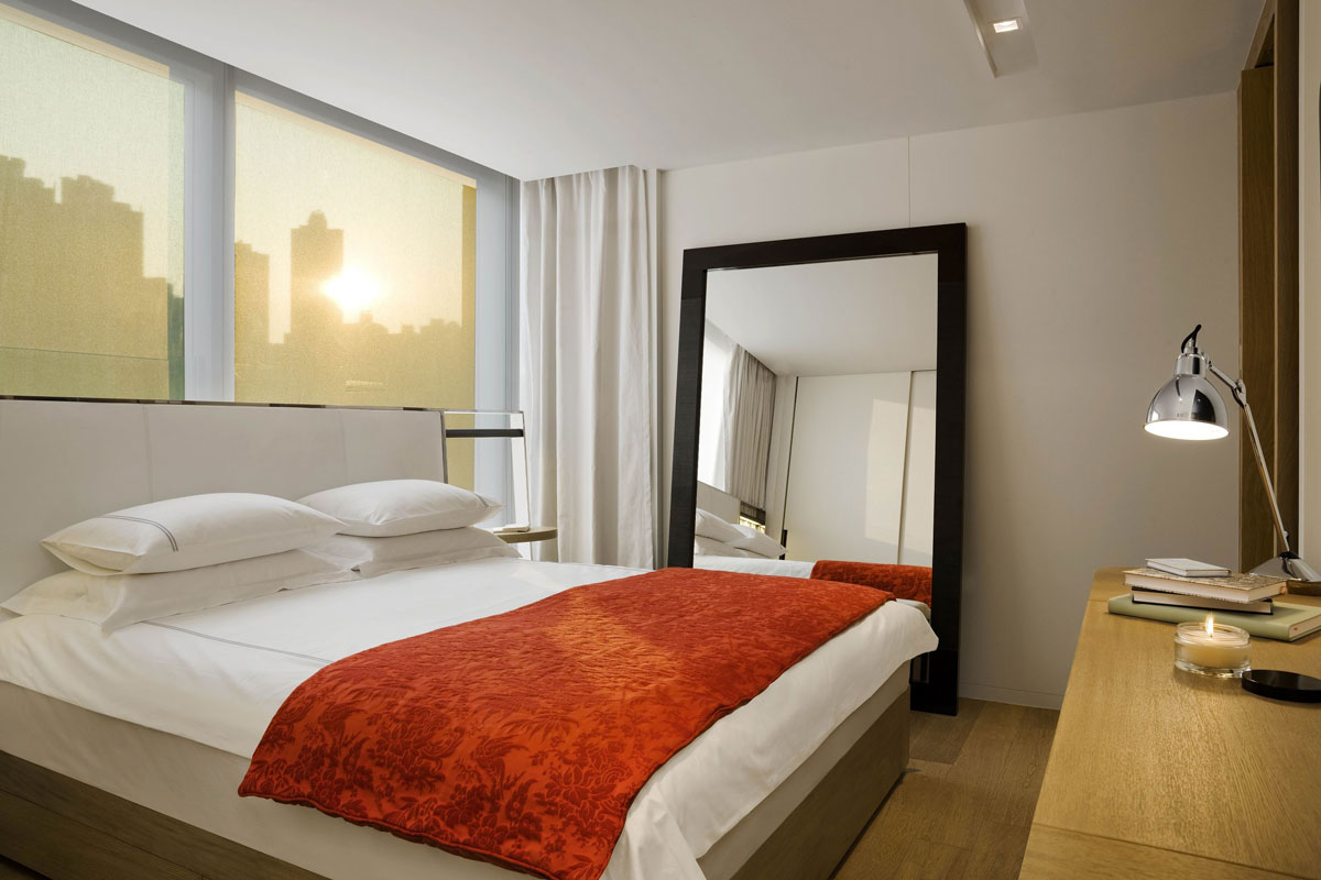 A spacious bedroom features a king-sized bed dressed in crisp white linens, accompanied by an orange accent blanket. Large windows allow soft, warm light to enter, illuminating the contemporary furnishings, including a full-length mirror and a wooden bedside table with a candle.