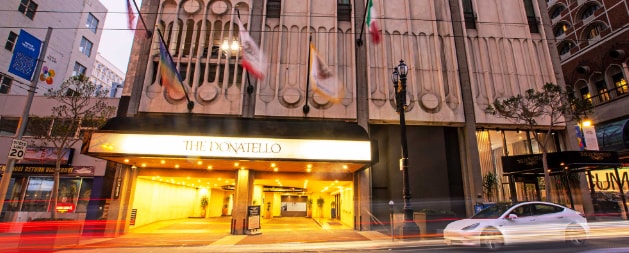 The entrance to The Donatello is depicted, featuring a spacious awning and illuminated signage. The building's architectural design includes distinctive textures and patterns. Flags are displayed prominently, and a vehicle is seen passing by in the foreground, highlighting the lively urban environment.