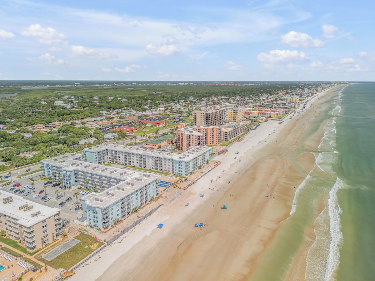 An aerial view captures the coastline of New Smyrna Beach, highlighting a wide stretch of sandy shore alongside several multi-story buildings. The clear blue sky is dotted with soft clouds, while the ocean gently meets the beach, creating a serene atmosphere.