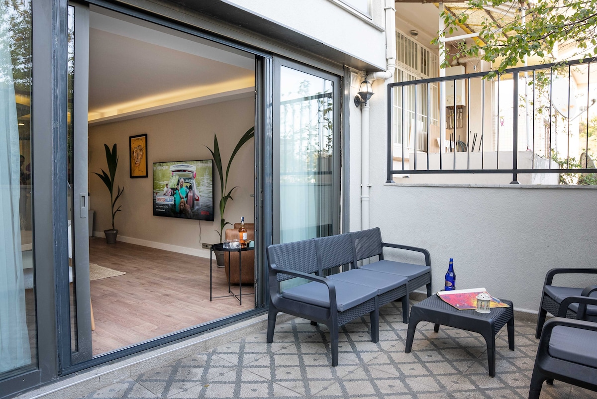 An inviting outdoor seating area features a two-person sofa and a small coffee table, offering a relaxed spot to enjoy the surroundings. Sliding glass doors lead into a spacious living area, with a large television visible in the background.