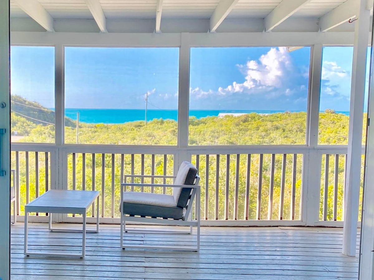 A screened-in porch features a light wooden floor and white railings, offering views of the turquoise ocean and lush greenery. A small table and a single chair provide a serene space for relaxation, framed by expansive windows that invite natural light inside.