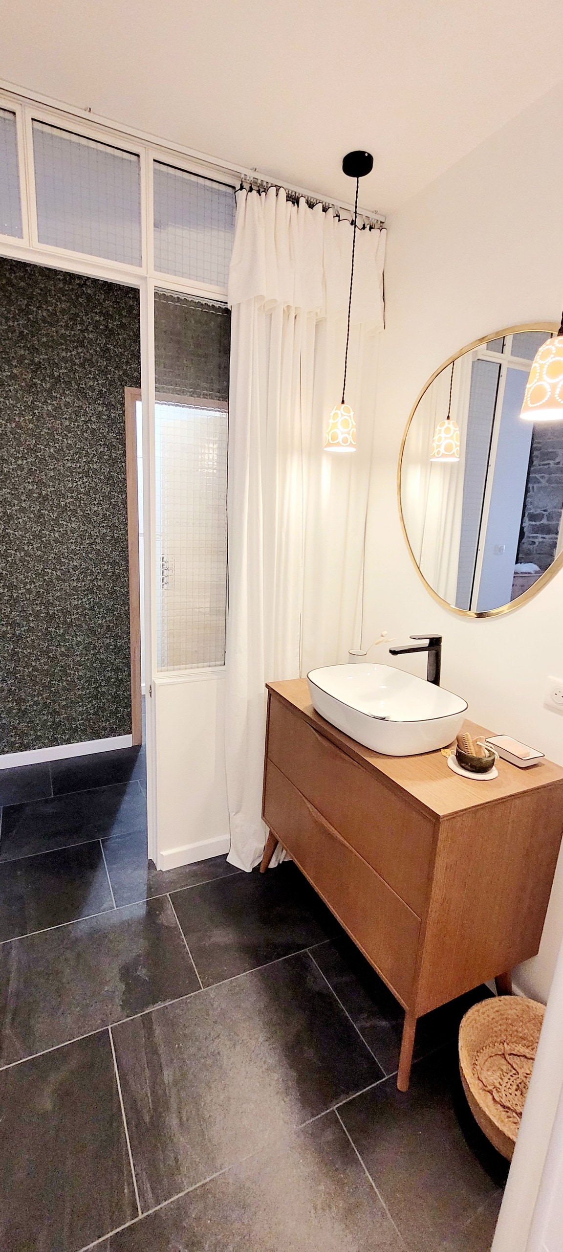 A modern bathroom is presented with a round mirror above a wooden vanity. A sleek faucet stands beside a white basin, while soft lighting is enhanced by two pendant lamps. Natural elements are suggested with a woven basket, and a partially obscured glass wall adds a subtle division.