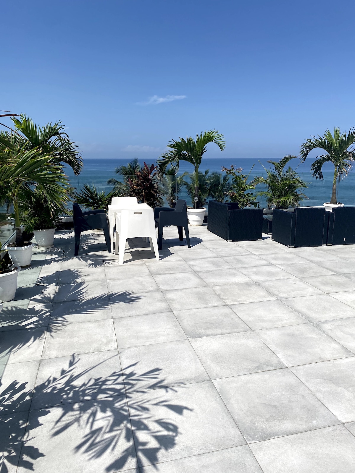 A designated outdoor seating area is visible on the spacious rooftop deck, featuring lounge chairs and a white table surrounded by tropical plants. The expansive ocean view provides a tranquil backdrop under clear blue skies.