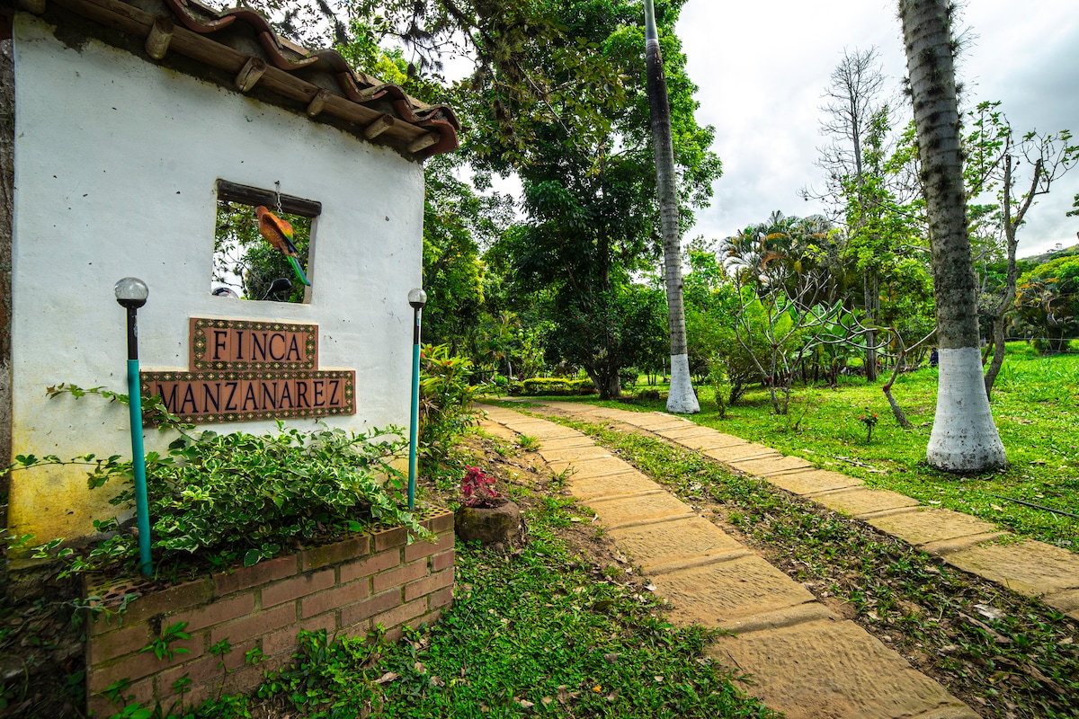 The entrance of Finca Manzanares features a rustic sign set against a backdrop of lush greenery. A stone path leads visitors through the well-kept gardens, surrounded by trees and vibrant foliage, inviting exploration of the tranquil surroundings.