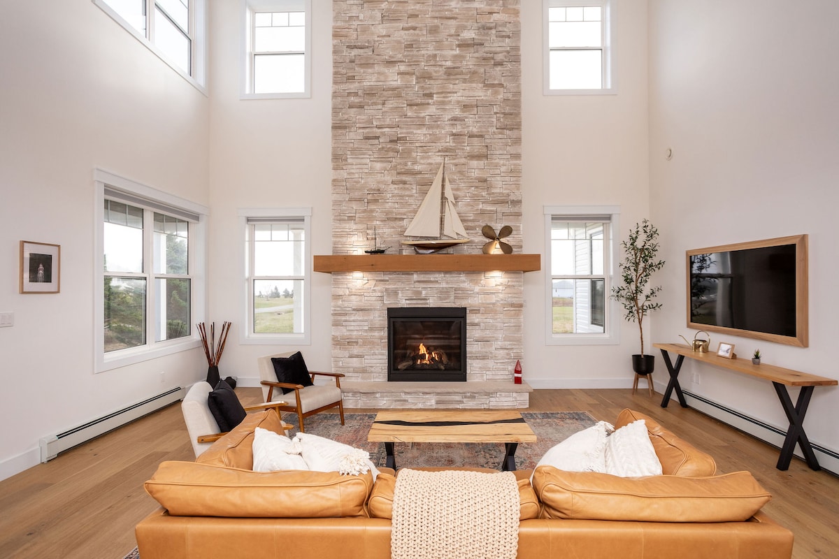 A spacious living area features a floor-to-ceiling stone fireplace as its focal point, adorned with decorative nautical elements. Large windows allow natural light to fill the space, with cozy seating arranged around a coffee table. A smart TV is mounted on the wall opposite the fireplace.