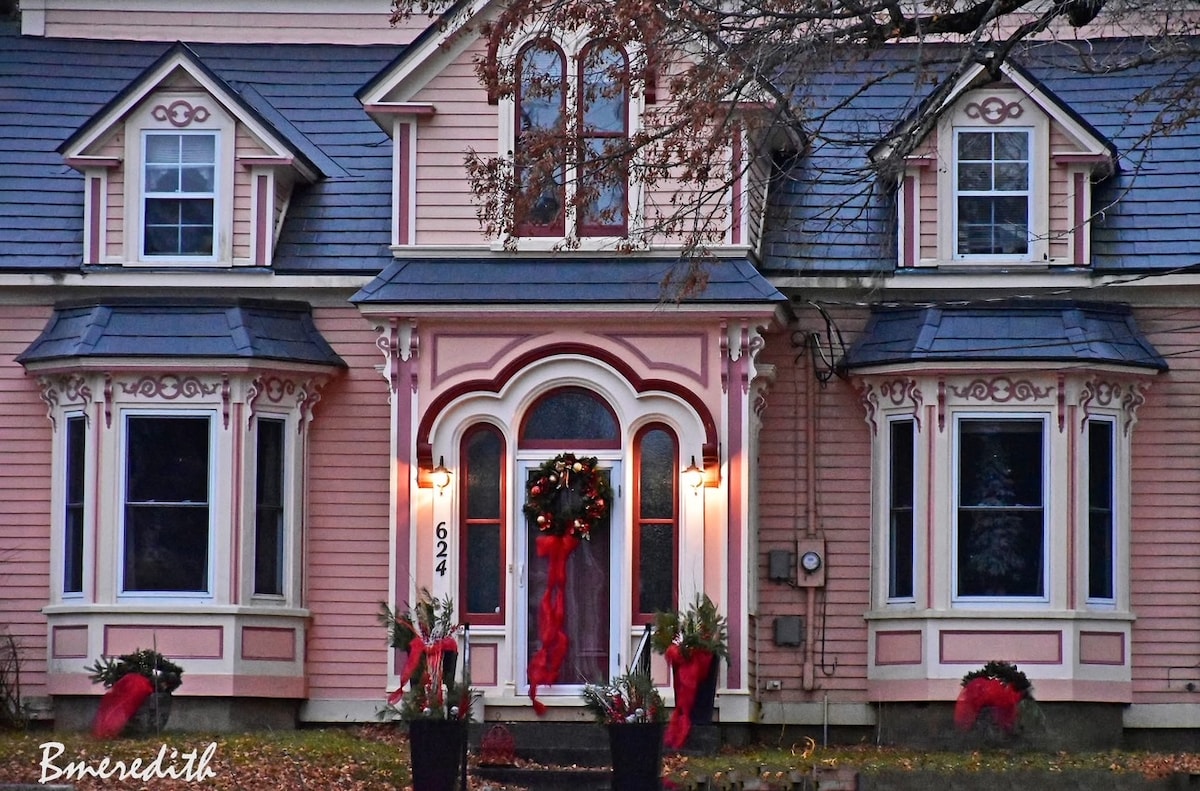 The charming exterior of Miller House showcases a pink façade with intricate decorative trim. Large bay windows are framed by lush greenery, and a festive wreath adorns the front door, which is flanked by decorative planters. Subtle lighting highlights the architectural details.