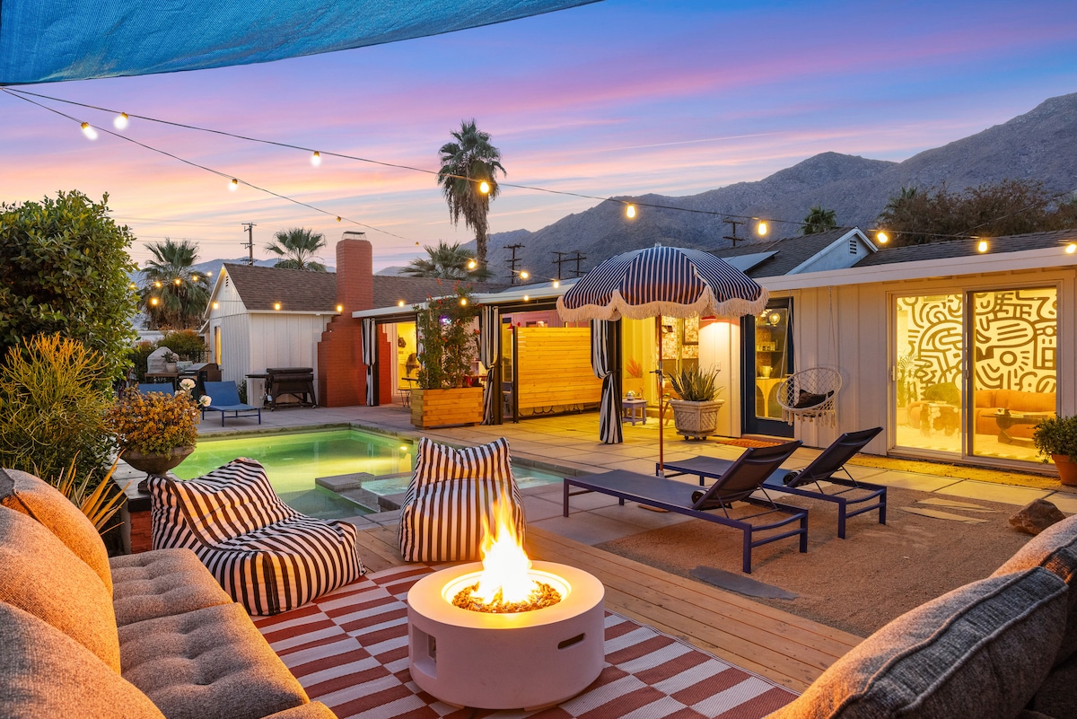 An inviting outdoor space features a cozy fire pit surrounded by patterned lounge chairs and umbrellas. The pool is visible, reflecting the colors of the sunset sky. String lights create a warm ambiance, while palm trees and mountains form a scenic backdrop.