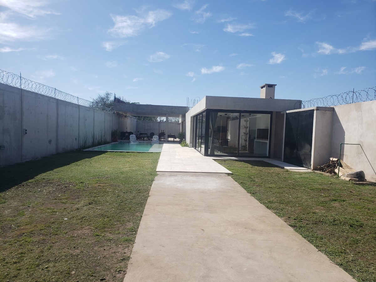 A sleek, modern structure is positioned beside a grassy lawn, leading to a serene pool area. A paved walkway extends towards the house, while a covered outdoor dining area and a surrounding perimeter wall can be seen in the background under a clear blue sky.