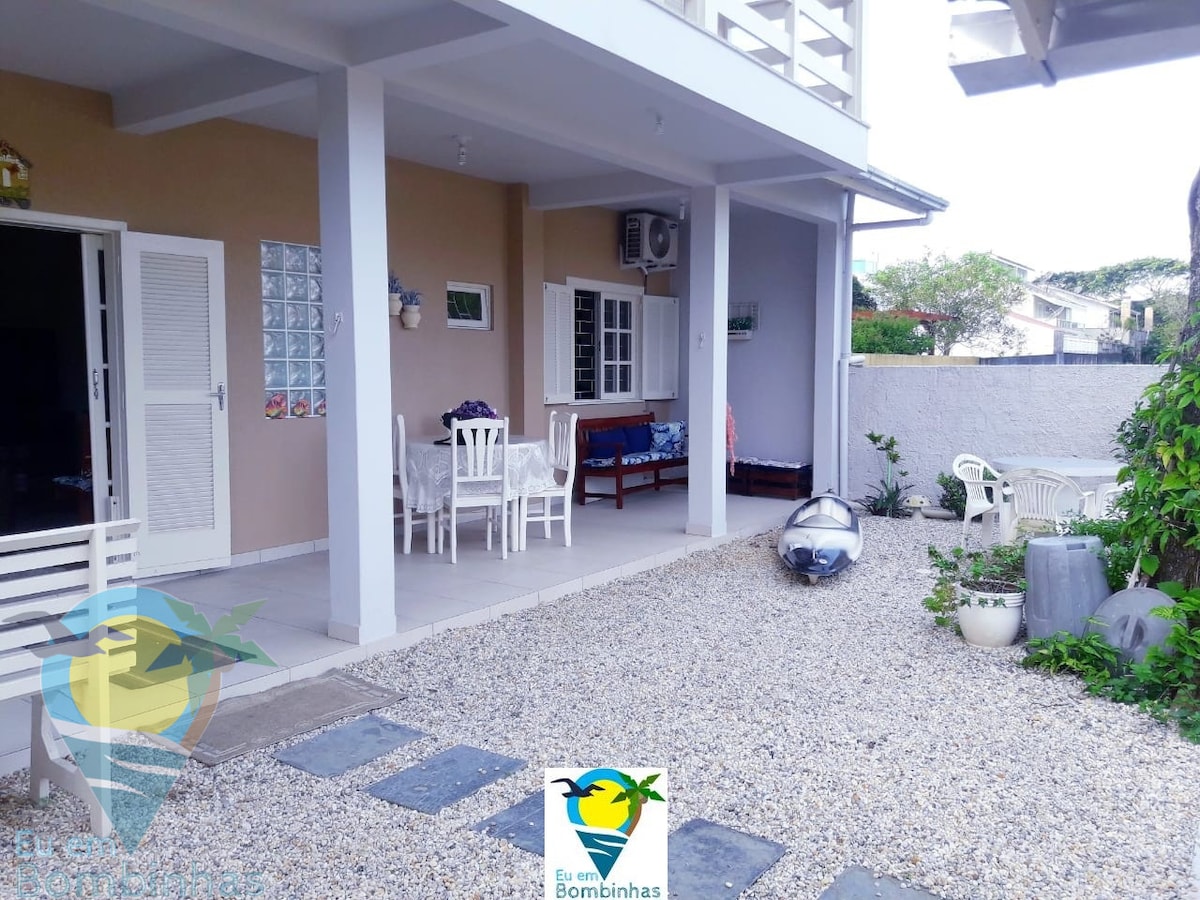 An outdoor area features a gravel surface with a small seating arrangement, including white chairs and a table for dining. A shaded porch highlights a sofa alongside a window, while ample space is provided for leisure activities.
