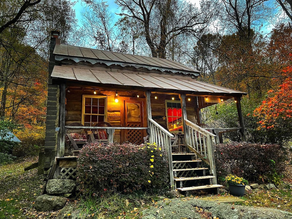 An antique log cabin is set amidst colorful autumn foliage, featuring a welcoming porch with wooden steps leading to the entrance. Soft lighting highlights the exterior, and an inviting array of seating is visible on the porch, enhancing the rustic charm of the cabin.