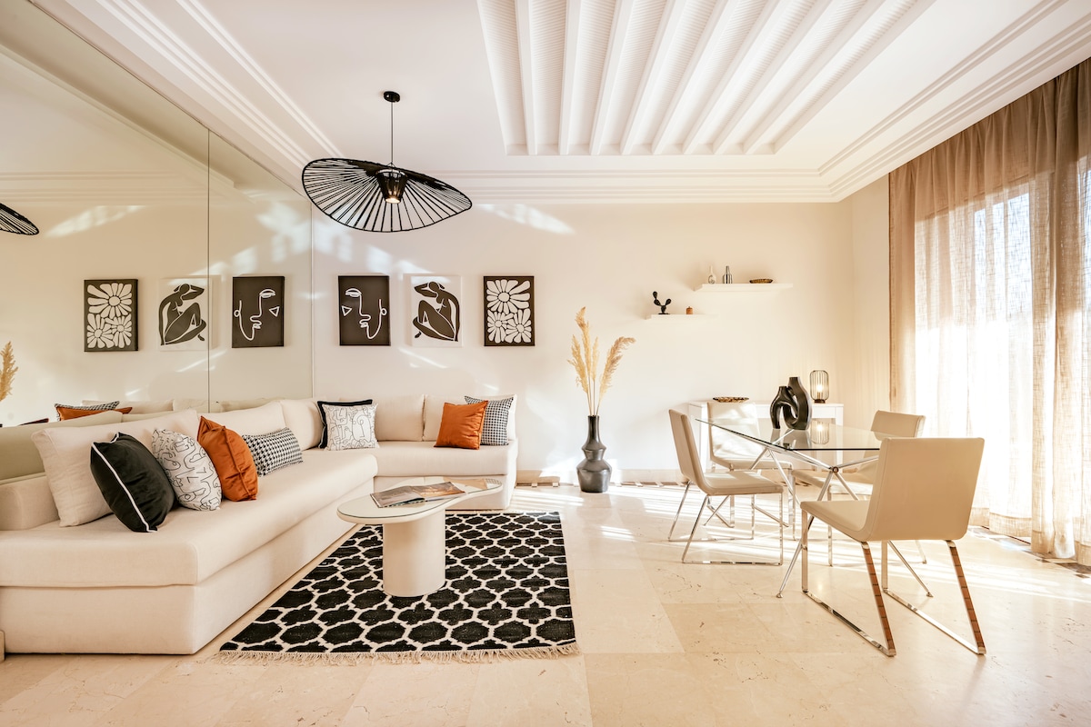 A light-filled living room features a spacious sofa adorned with decorative pillows in soft tones. A glass dining table with seating for multiple guests sits beside a patterned rug. Abstract artwork decorates the walls, and natural light streams through large windows adorned with sheer curtains.