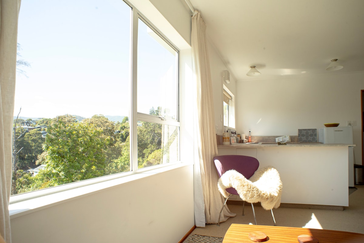 A bright and airy living space features large windows that allow natural light to fill the room. A purple chair with a textured throw sits near the window, with a small wooden table visible in the foreground. A kitchenette is partially seen in the background.