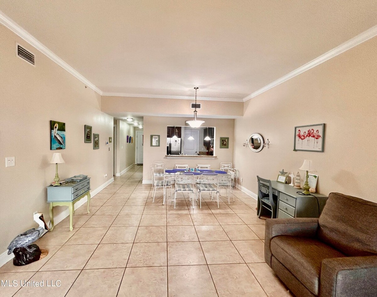 An open living space features tiled flooring and neutral-colored walls. A dining table with several chairs is positioned centrally, while a comfortable sofa sits to one side. Decorative artwork and accents are visible throughout the room, adding character to the inviting atmosphere.