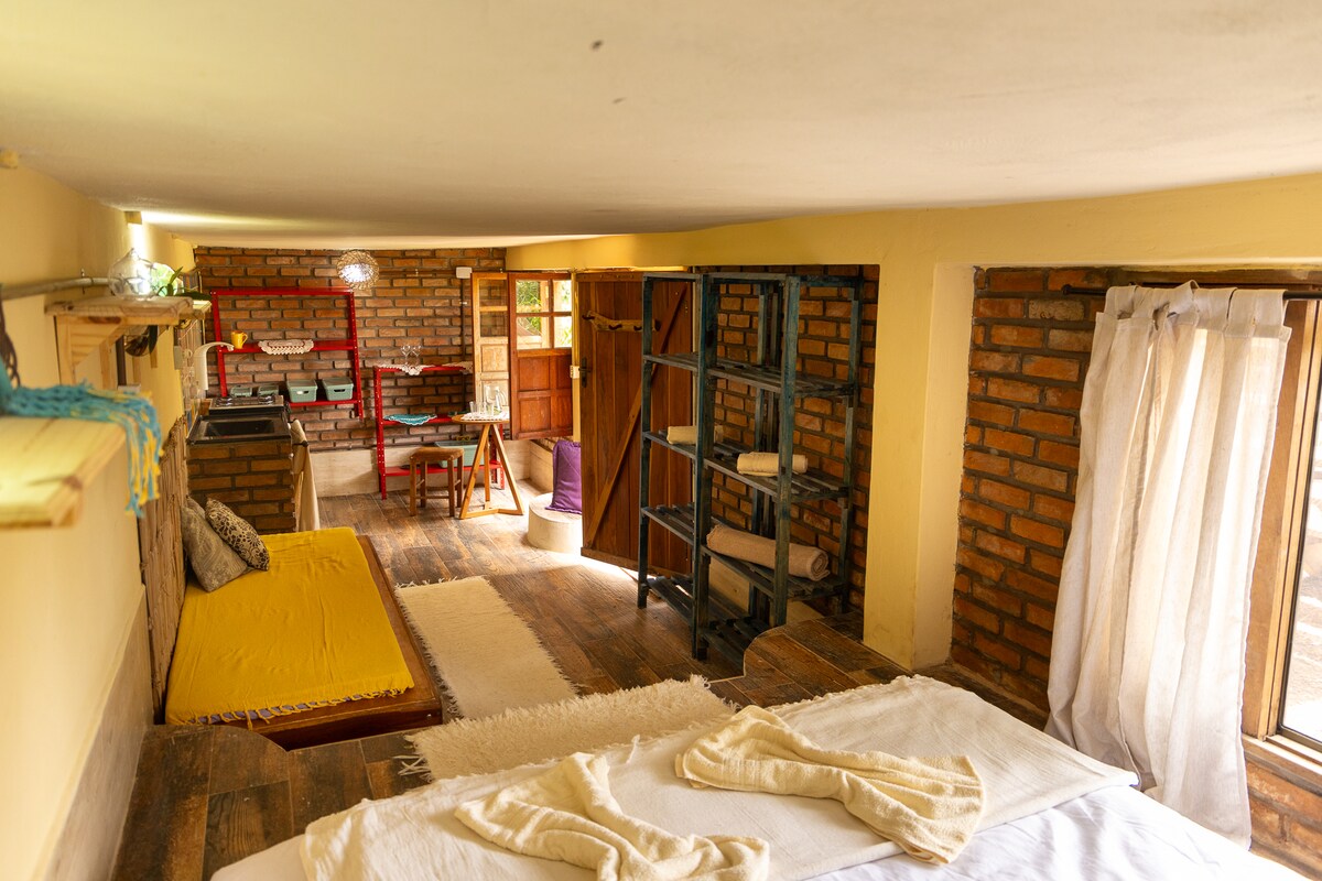 A cozy sleeping area features a spacious bed with soft linens and a light-colored blanket. Exposed brick walls frame the room, complemented by wooden floors. Natural light streams through two windows, illuminating the inviting decor, which includes a yellow accent area and various shelves.