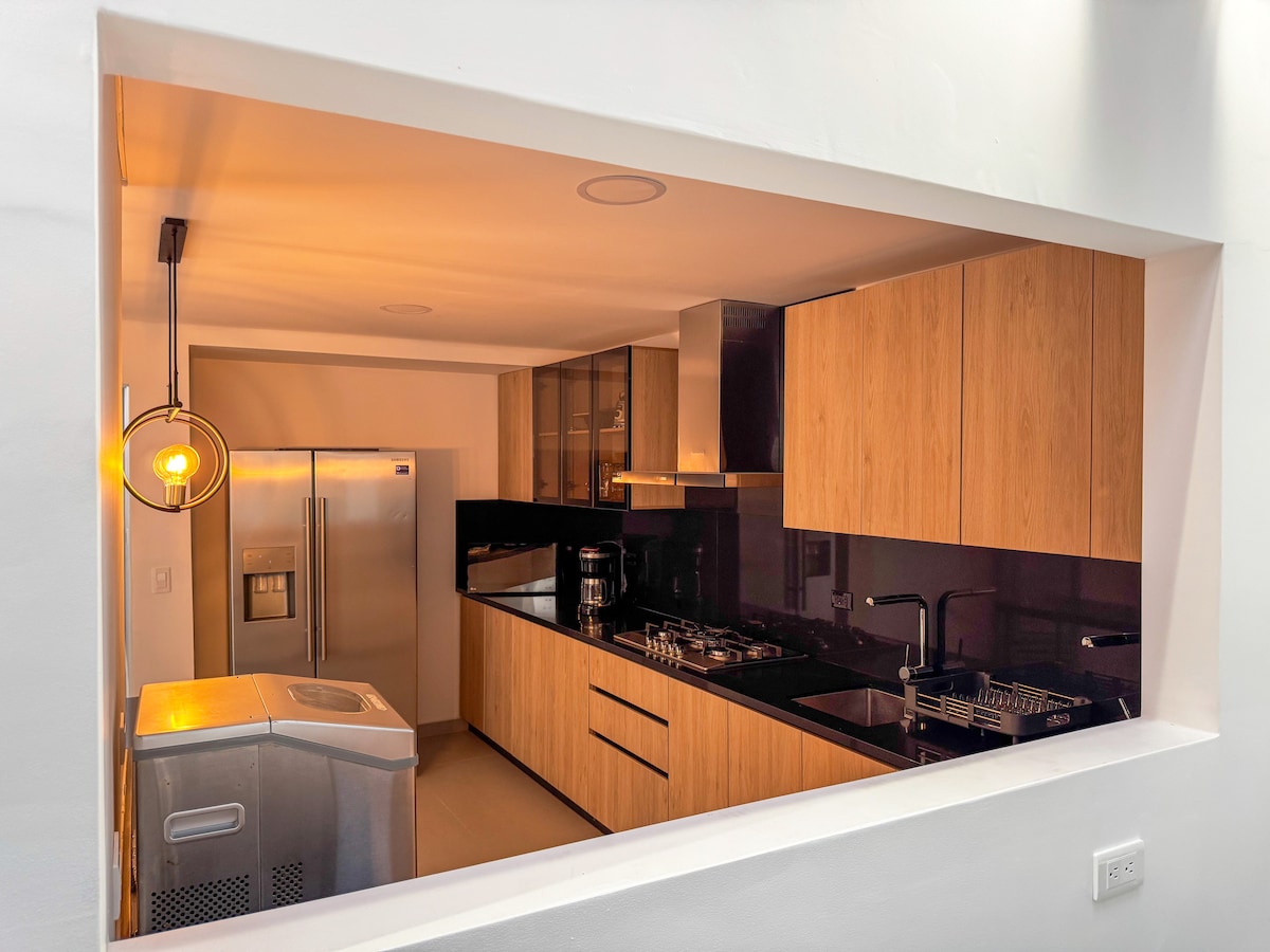 A modern kitchen with wood cabinetry and sleek black countertops is visible through an open wall. Stainless steel appliances, including a refrigerator and stove, are positioned for convenience. Soft lighting enhances the clean lines and spacious feel of the area.
