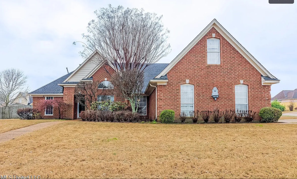 The exterior of a large brick home is featured, set against a clear sky. A neatly landscaped front yard is visible, with carefully trimmed shrubs and grass. Symmetrical windows and a front door add architectural interest, framed by leafy trees in the background.