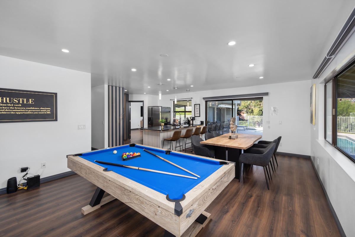 A spacious open living area features a vibrant blue pool table at its center, surrounded by comfortable seating arrangements. Large windows offer visibility to the outdoor pool area, while a modern kitchen and dining space are evident in the background.