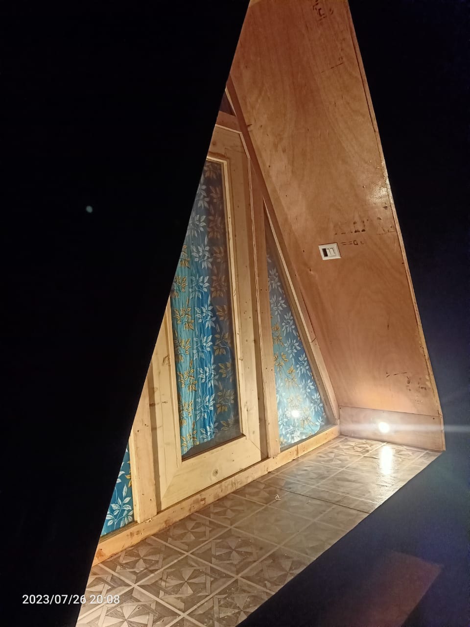 The interior of the A-frame cabin features wood-paneled walls and a brightly patterned window curtain. Soft lighting illuminates the space, highlighting the unique architectural angles. A textured floor completes the cozy atmosphere, inviting relaxation.