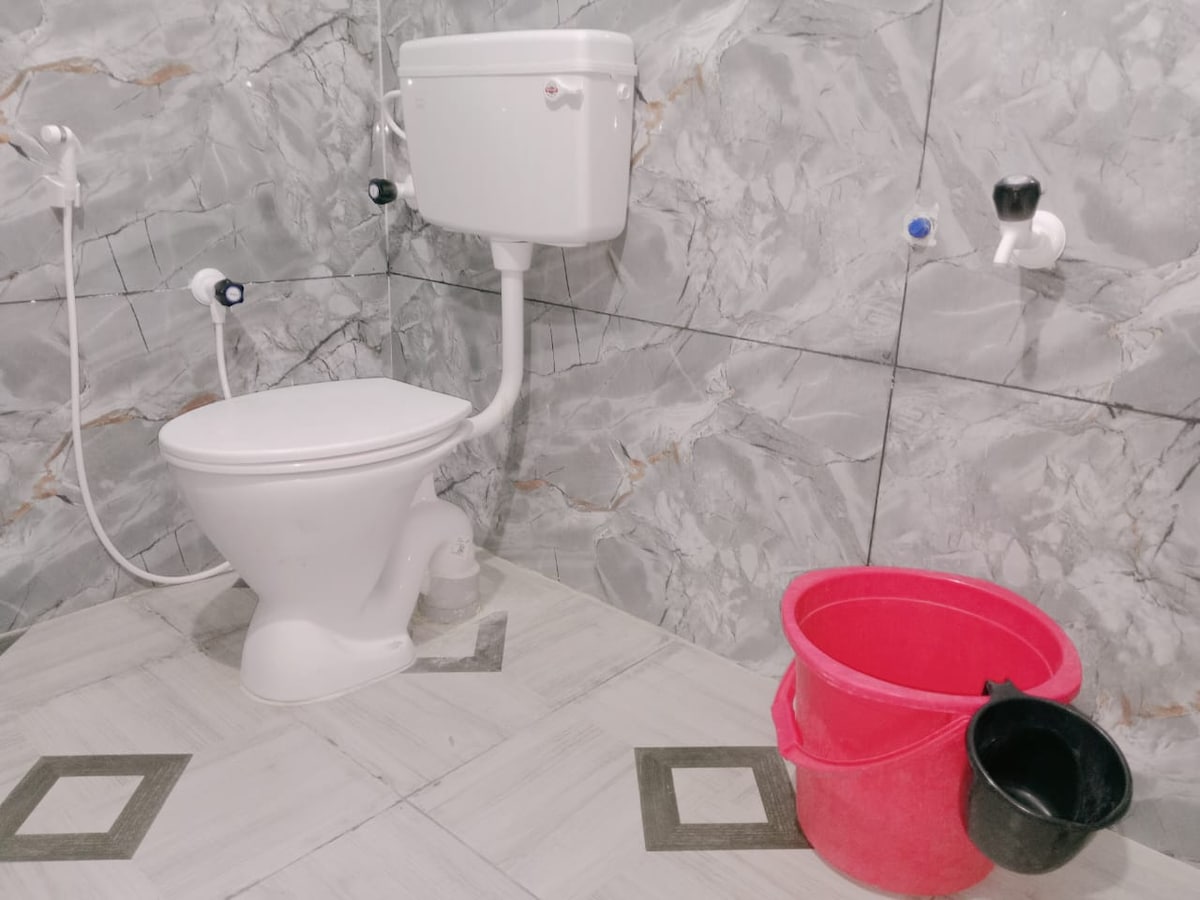 A clean bathroom area is featured with modern marble-patterned walls and floor tiles. A white toilet is positioned against the wall, alongside a pink bucket and a black bucket near the floor. Plumbing fixtures are visible on the wall.