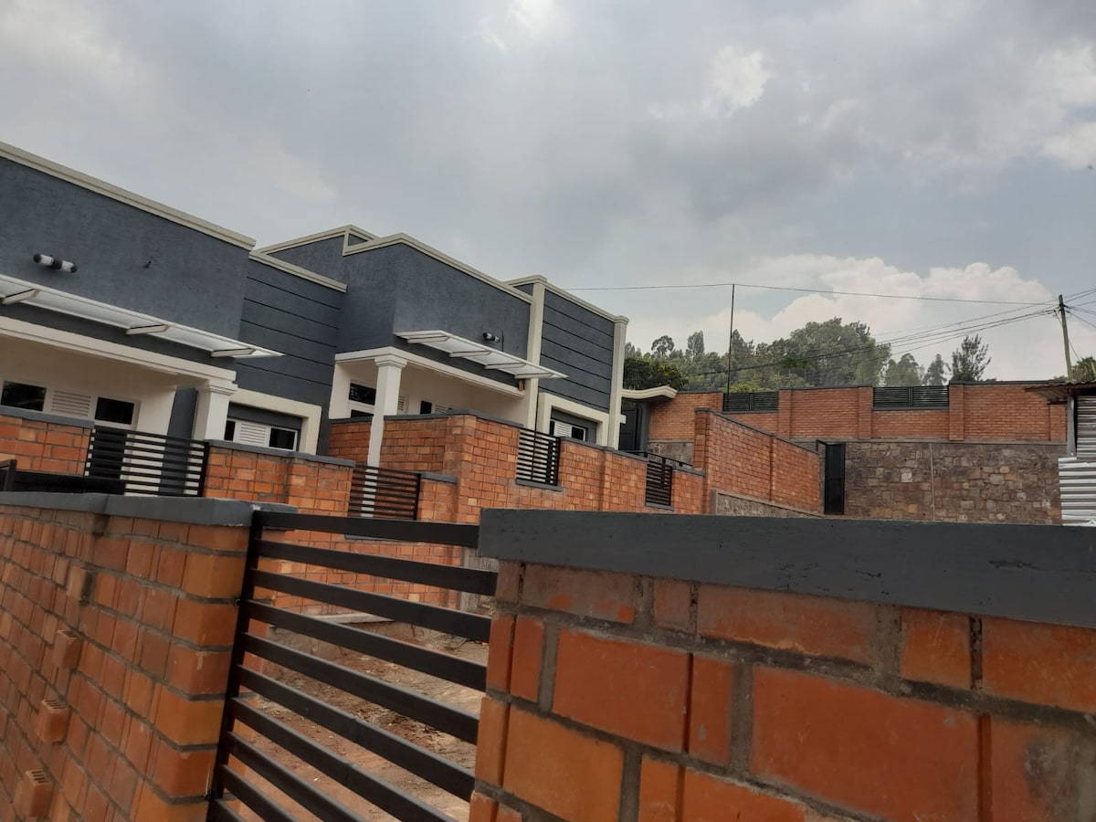 A modern two-story house is depicted, featuring brick walls and a gray exterior. The property is enclosed by a secure fence, providing privacy. Surrounding areas consist of additional brick structures and greenery, with a cloudy sky above, enhancing the serene environment.