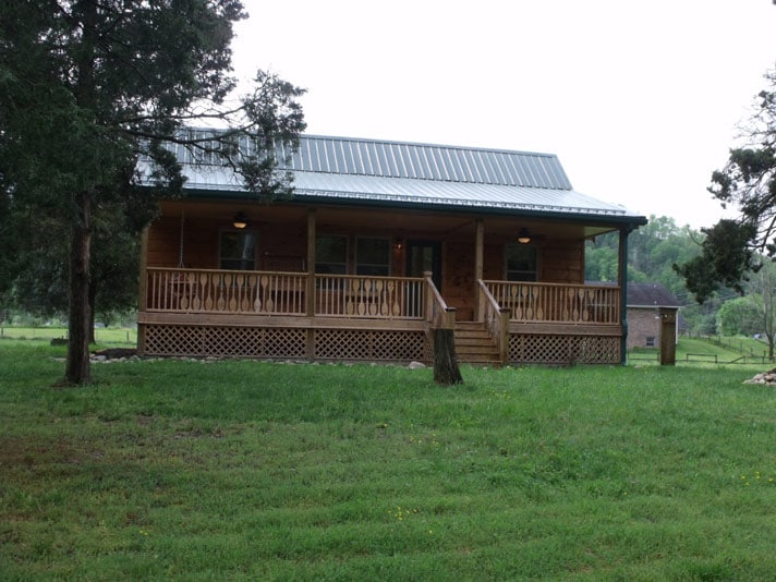 A rustic cabin with a wooden façade is shown, featuring a porch with railings and overhead lighting. Surrounding green grass and trees create a natural environment. A gravel walkway leads to the entrance, enhancing the cabin's accessibility to guests.