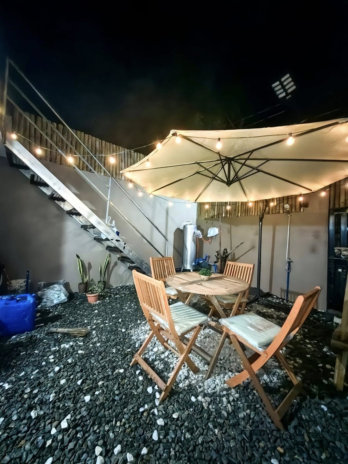 A cozy outdoor dining area is highlighted by a spacious table surrounded by four wooden chairs. An umbrella provides shade, while string lights create a soft glow. A pebble surface complements the setting, and stairs are visible in the background, leading to an upper level.