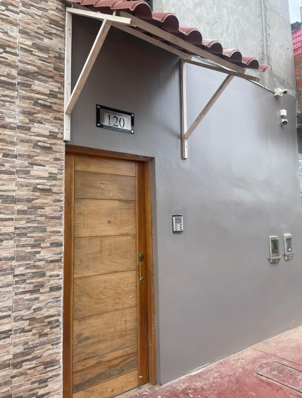 The entrance of the building is showcased, featuring a wooden door with natural grain and a number 120 displayed above. The exterior walls are painted in muted grey with a decorative stone texture, complemented by a red-tiled awning overhead.