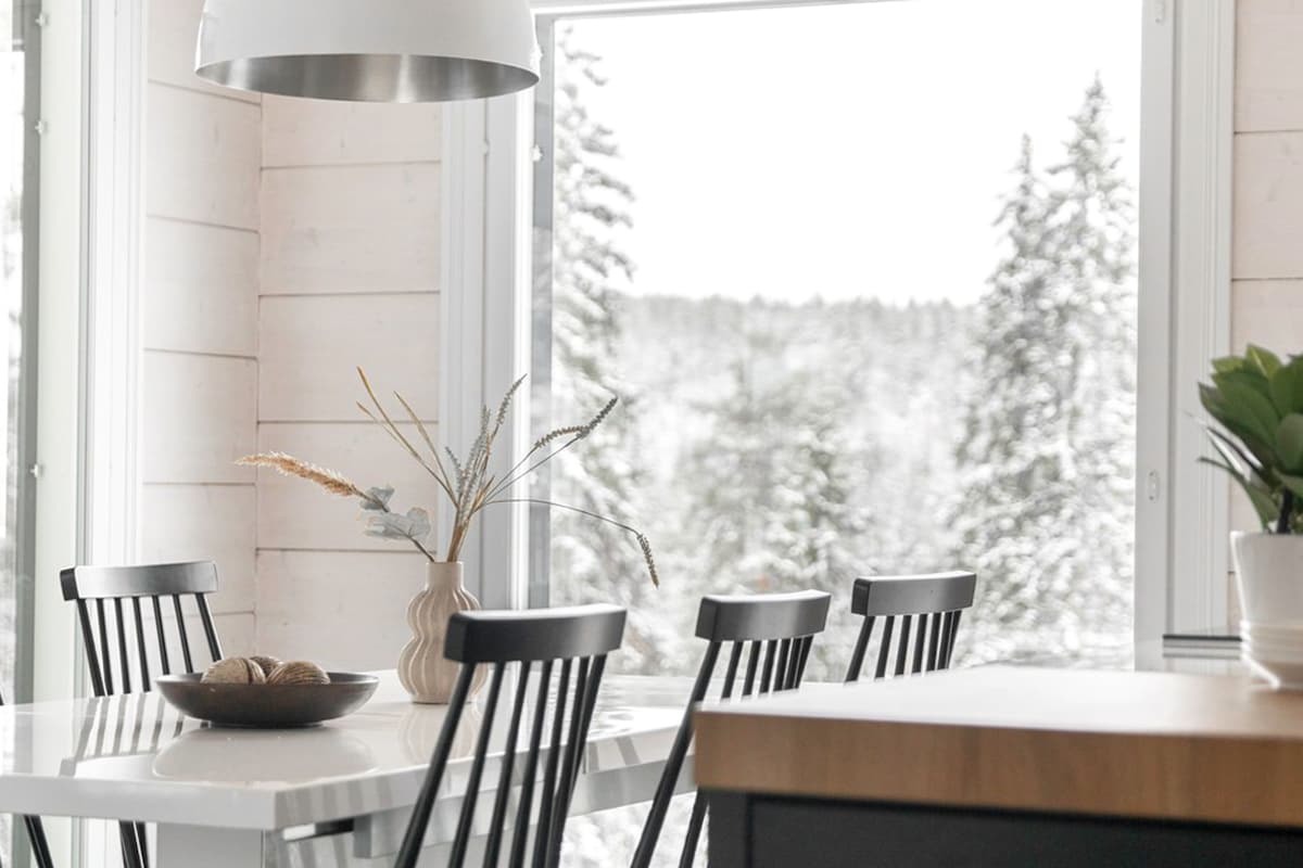 A bright dining area features a white table surrounded by black chairs, enhancing the sense of space. Large windows provide views of a snowy landscape, while minimalistic decor includes a vase with dried flowers and a bowl with natural elements.