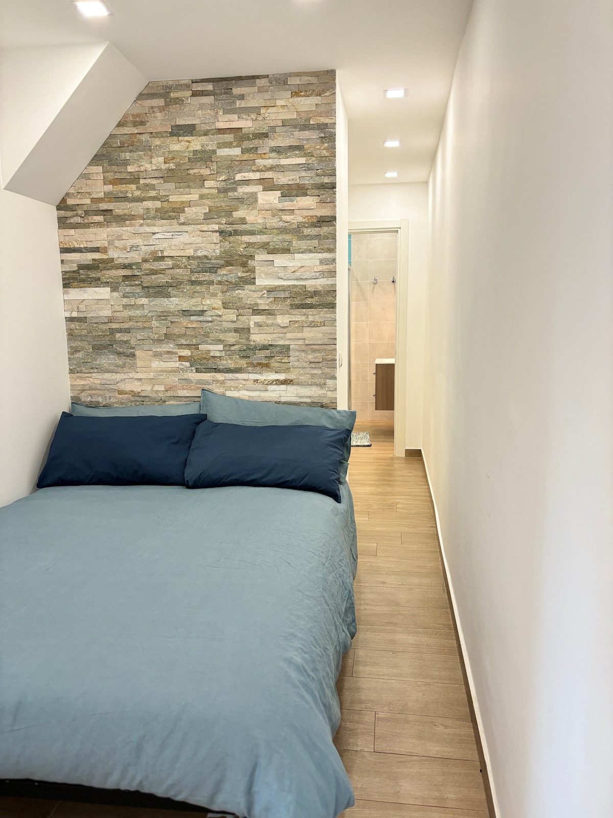 A compact sleeping area is highlighted by a single bed covered in a soft blue duvet. The wall features a stone veneer design, adding texture to the space. A doorway at the end leads to a bathroom area, with neutral tiles visible in the open entry.