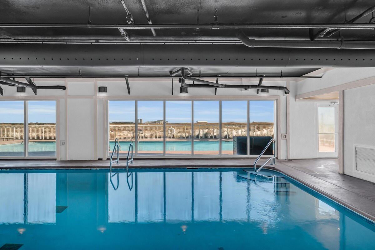 An indoor swimming pool is visible with clear blue water. Large windows allow natural light to fill the space, showcasing views of the outdoor pool and landscape beyond. Poolside seating can be seen in the background, inviting relaxation.