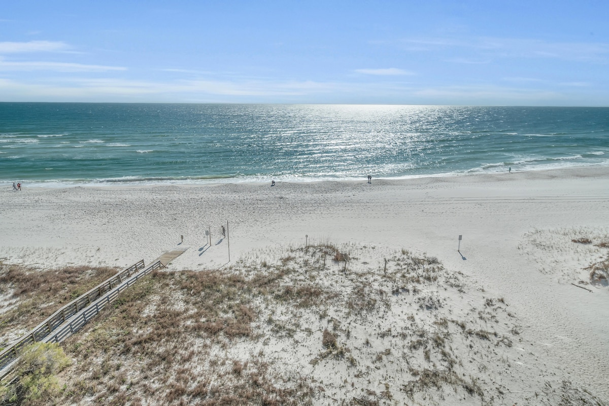 A serene view of the Gulf of Mexico unfolds, showcasing soft white sands and gentle waves under a clear sky. The beach is sparsely populated with individuals walking along the shore, creating a tranquil coastal atmosphere.