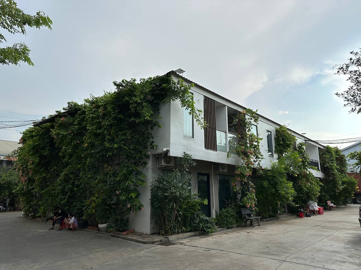 An exterior view of a two-story building is surrounded by lush greenery, with vines climbing the facade. Large windows allow ample natural light into the spaces. The entrance is framed by a variety of plants, enhancing the tranquil ambiance of the setting.