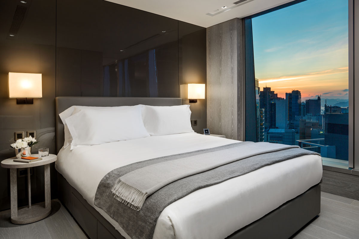 A spacious bed is positioned against a sleek dark feature wall, illuminated by soft bedside lamps. A grey throw drapes over the bedding, while floor-to-ceiling windows offer a stunning view of the city skyline against a sunset backdrop.