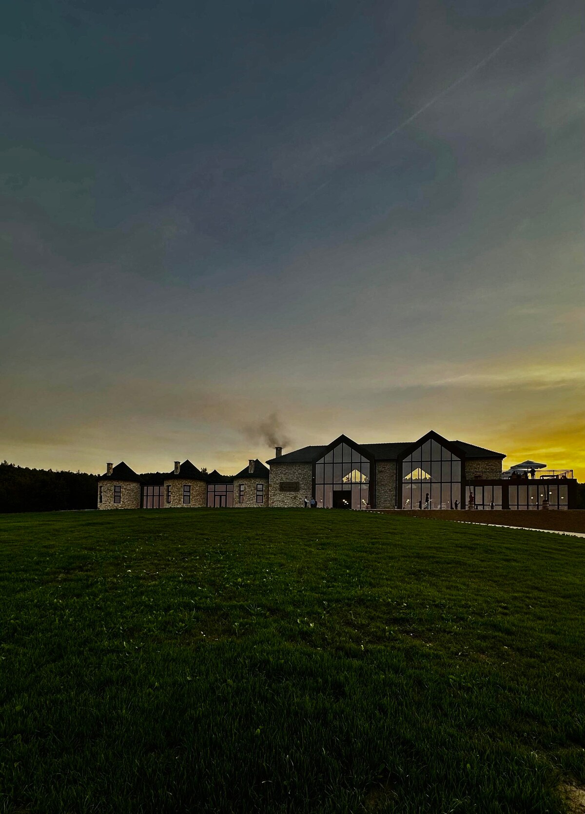 A spacious building stands on a gently sloping green landscape, with large windows reflecting the soft colors of the evening sky. The structure features a combination of architectural styles, evident in its multiple rooftops and stone facade, creating an inviting silhouette against the horizon.