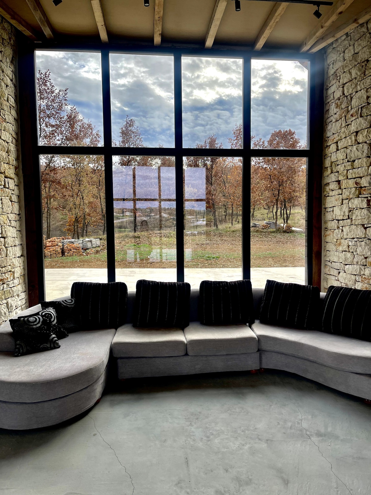 A circular gray sofa is positioned in front of a large window, providing a view of a serene outdoor landscape with trees showing autumn foliage. The room features stone walls and a polished concrete floor, creating a cozy yet modern ambiance.