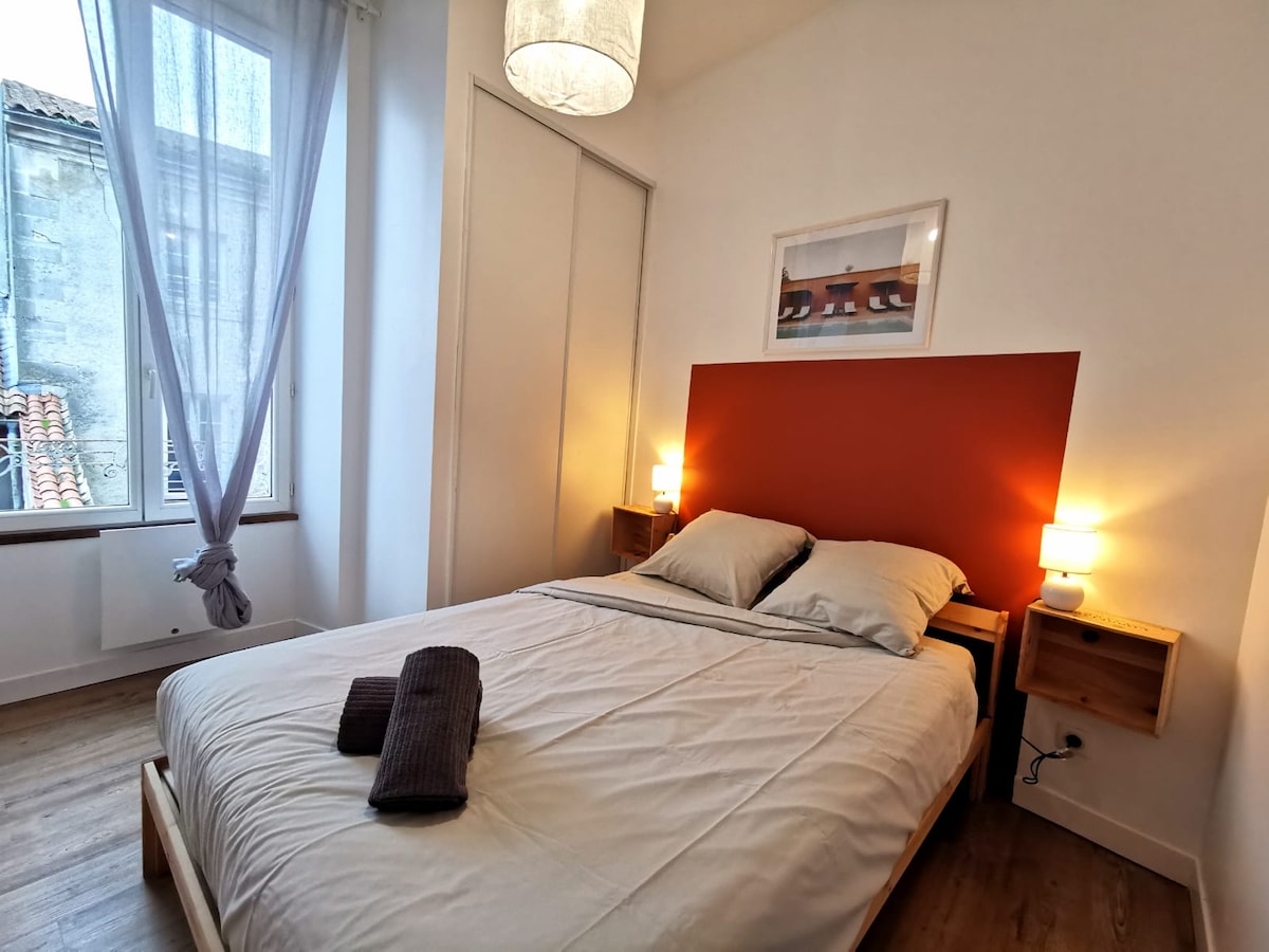 A bright bedroom features a spacious bed with light linens, complemented by two bedside tables with lamps. Natural light filters in through a window adorned with sheer curtains, while a simple piece of artwork is displayed above the headboard.