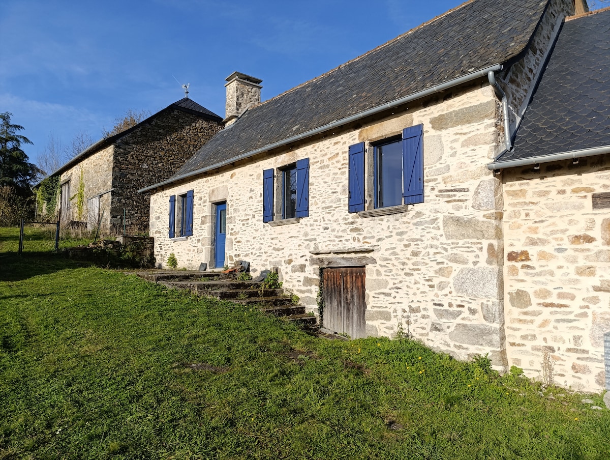 A rustic farmhouse exterior is presented, showcasing textured stone walls and blue shutters. Surrounding greenery enhances the natural setting, with steps leading to the front door. The simplicity of the landscape and architecture reflects a peaceful countryside atmosphere.
