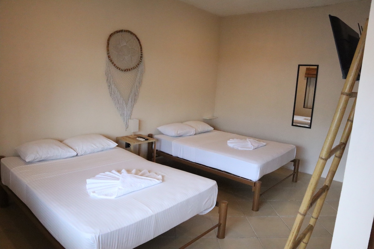 A serene sleeping area featuring two spacious beds with crisp white linens. Natural light illuminates the room, highlighting a decorative wall hanging and a slim mirror. A small wooden nightstand is positioned beside one bed, while a contemporary TV is mounted on the opposite wall.