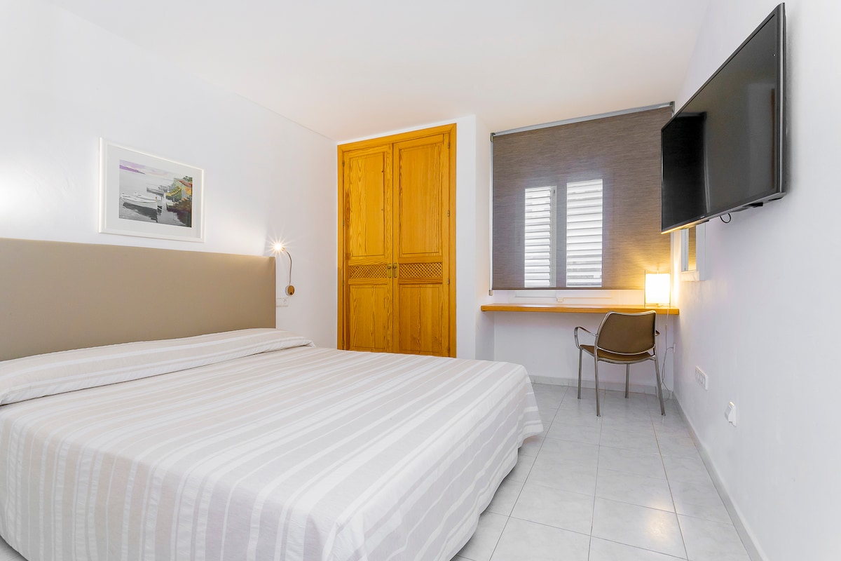 A well-lit bedroom features a spacious bed with light-colored linens. A wooden wardrobe stands against one wall, while a desk and chair are positioned near a window. The room is complemented by a mounted television and minimalistic decor.