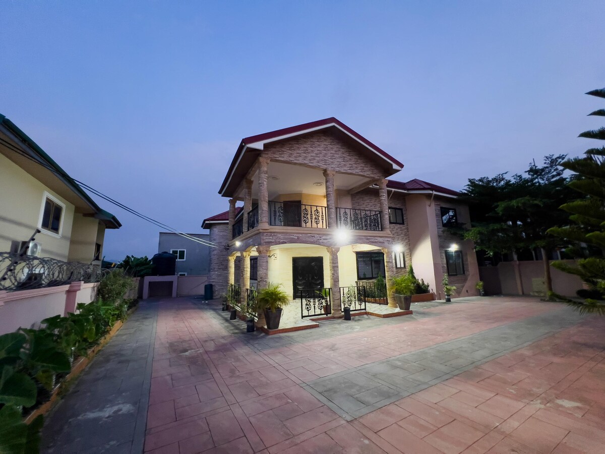 The two-story house features a welcoming facade with a combination of textured stone and smooth plaster. Multiple windows allow for natural light, while balcony areas provide outdoor views. Surrounding the house, a paved driveway is complemented by landscaped plants and trees.