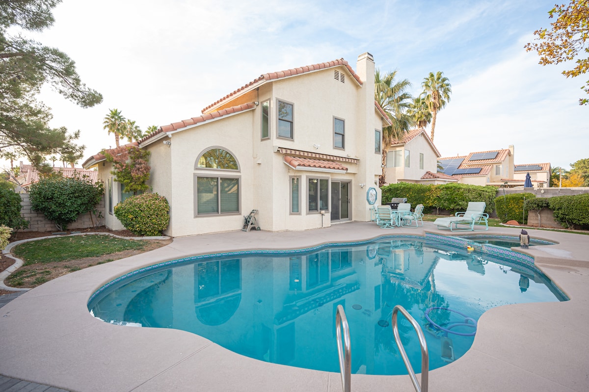 A two-story home with a charming exterior is depicted, featuring a sparkling pool surrounded by a spacious patio. Comfortable lounge chairs are situated nearby, while palm trees and well-maintained hedges provide a serene backdrop.