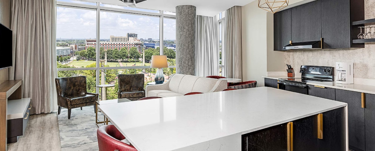 An open living space is showcased with large windows revealing views of the city landscape. A white sofa and two armchairs are arranged around a coffee table. A stark white kitchen island with black accents is visible, adding to the contemporary design.