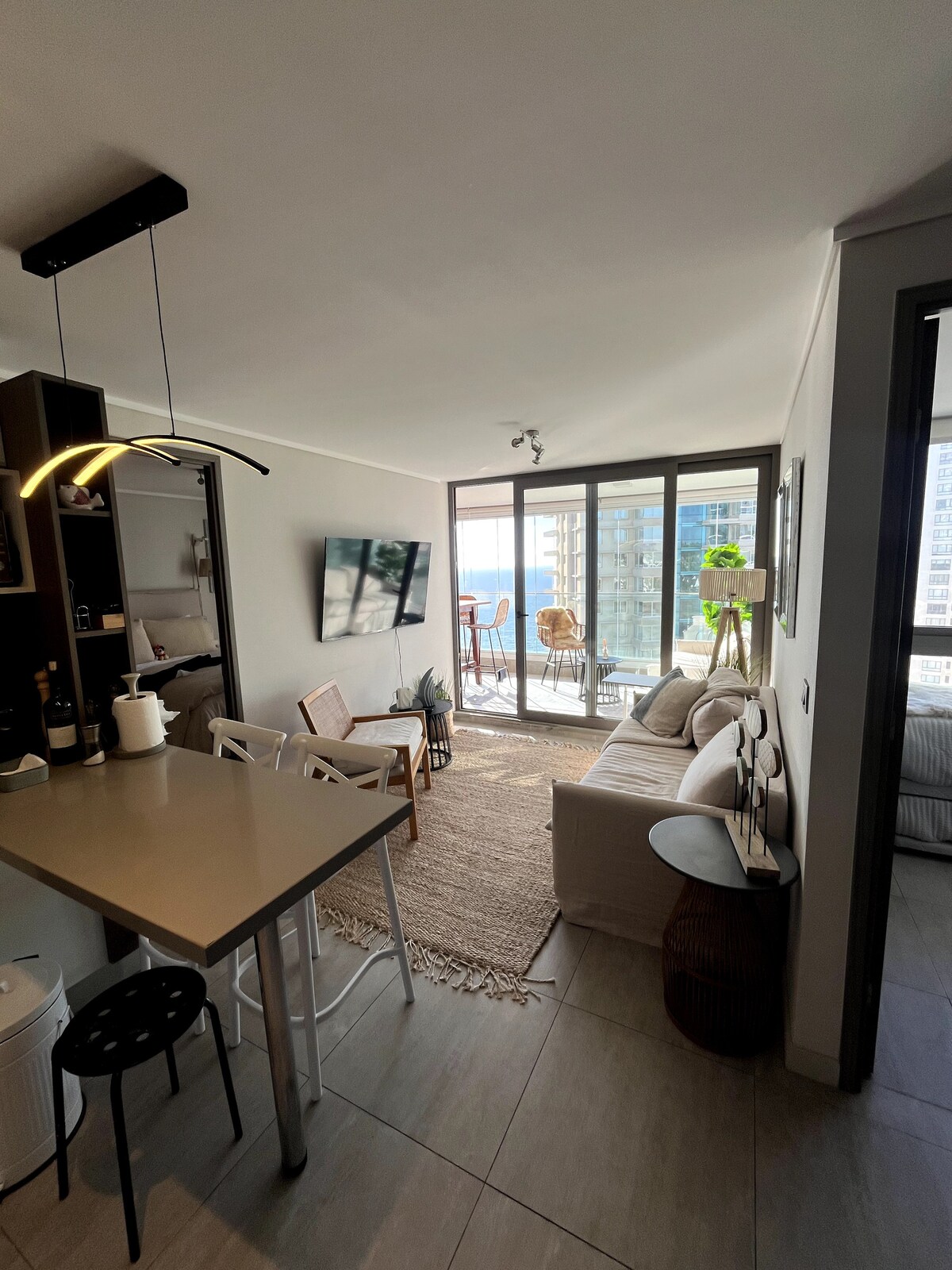 A modern living area is presented with large windows allowing natural light to fill the space. A light-colored sofa sits on a woven rug, complemented by a dining table and stylish bar stools. A small balcony is visible, featuring outdoor seating and city views.