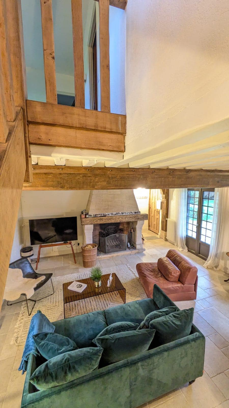 A spacious living area is highlighted by a functional fireplace and large windows offering natural light. Comfortable seating includes a plush green sofa and a coral-colored armchair, arranged around a low coffee table. A TV is positioned beside a staircase leading to the upper level.