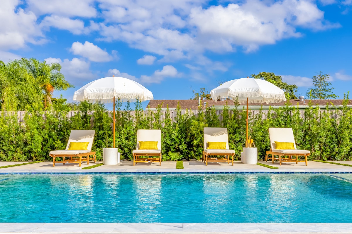 A private pool area is showcased with three sun loungers positioned beneath elegant white umbrellas. Lush greenery surrounds the pool, providing a serene backdrop against a clear blue sky, creating an inviting space for relaxation and enjoyment.